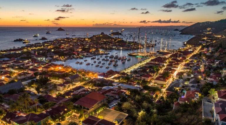 Aerial view of a coastal town at sunset with a busy harbor full of sailboats and waterfront housesPalm lined streets glow with lights along the shore