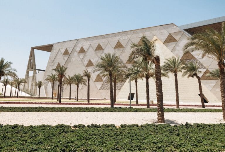 Geometric beige building with triangular patterns flanked by palm trees and desert landscaping