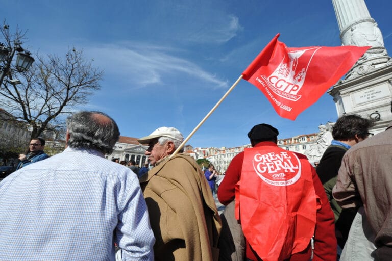 Protesters gather in a city square with a raised red CGTP flag a person wears a red Greve Geral vest near a white monument against a blue sky