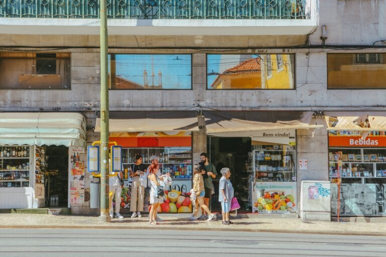 People stand and chat in front of a row of shops on a sunny street with fruit posters visible in the storefront windows above them