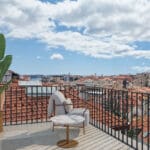 Balcony with a marble round table gray cushioned chair a tall potted plant and a city of red tiled roofs beyond the railing under a blue sky