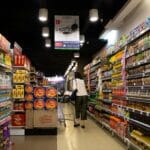 Shoppers navigate a supermarket aisle between tall shelves filled with colorful bottled drinks on both sides a person pushing a cart ahead under bright ceiling lights