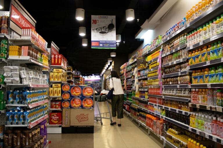 Shoppers navigate a supermarket aisle between tall shelves filled with colorful bottled drinks on both sides a person pushing a cart ahead under bright ceiling lights