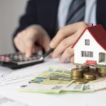 Small white house model with a red roof sits on stacks of coins beside a calculator and financial papers symbolizing real estate costs or mortgages