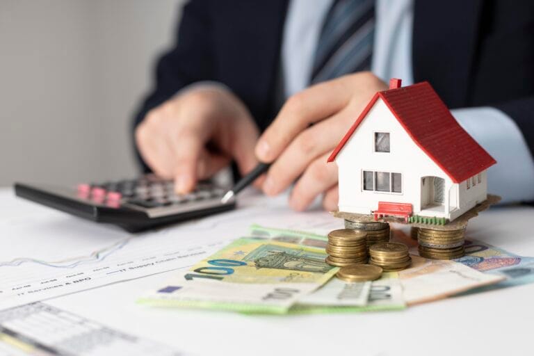 Small white house model with a red roof sits on stacks of coins beside a calculator and financial papers symbolizing real estate costs or mortgages