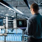 Man in a blue shirt stands in a data center looking at a laptop displaying a glowing network visualization
