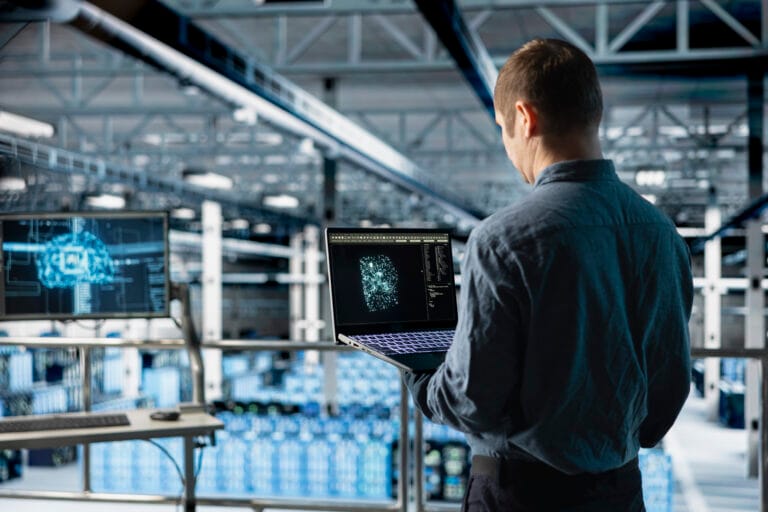 Man in a blue shirt stands in a data center looking at a laptop displaying a glowing network visualization