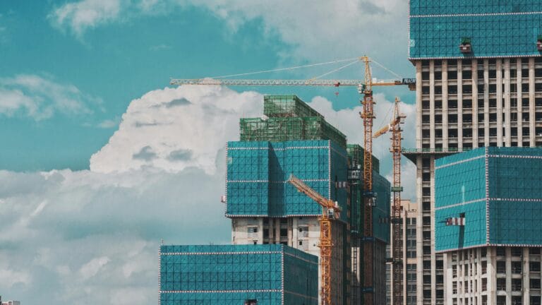 Construction site with tall buildings wrapped in blue green safety netting and multiple cranes overhead against a cloudy sky