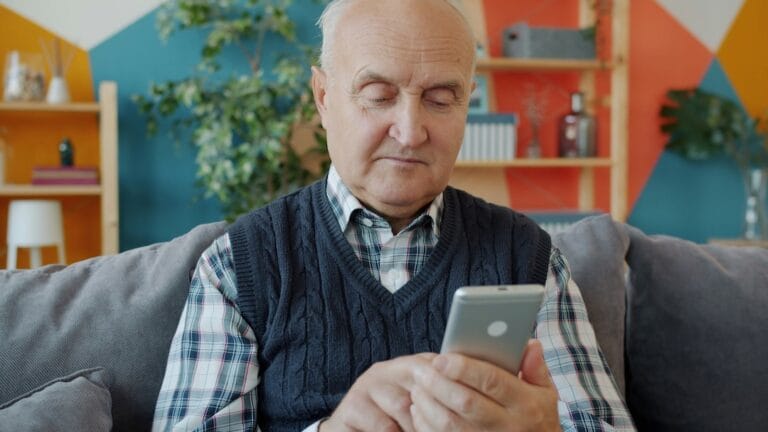 Older man on a gray couch looking at his smartphone in a colorful living room