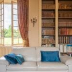 Bright living room with an open window ornate gold curtains a white sofa with blue pillows and a tall bookshelf filled with books outside the window view of greenery