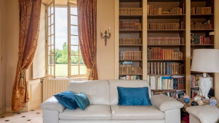 Bright living room with an open window ornate gold curtains a white sofa with blue pillows and a tall bookshelf filled with books outside the window view of greenery