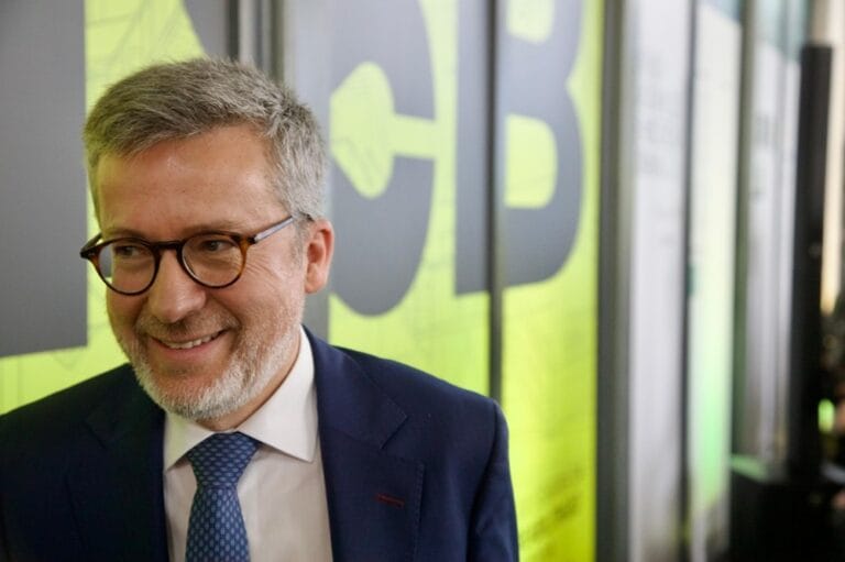 Smiling man in a navy suit and blue tie wearing round glasses standing in front of a lime green wall with large letters behind him