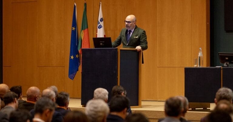A man in a suit gives a speech from a podium on a stage with multiple flags behind him and an audience in front