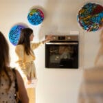 Woman in a pale yellow suit reaches to operate a built in oven in a room with colorful circular wall art and onlookers nearby