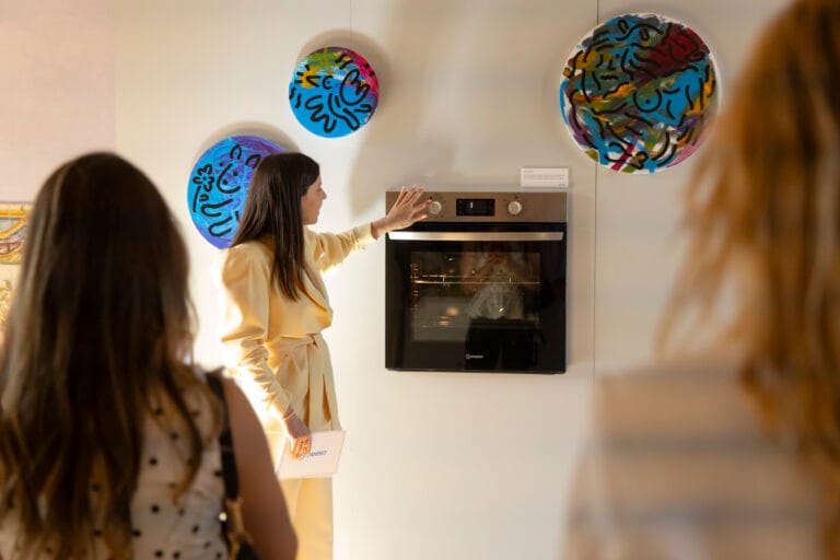 Woman in a pale yellow suit reaches to operate a built in oven in a room with colorful circular wall art and onlookers nearby