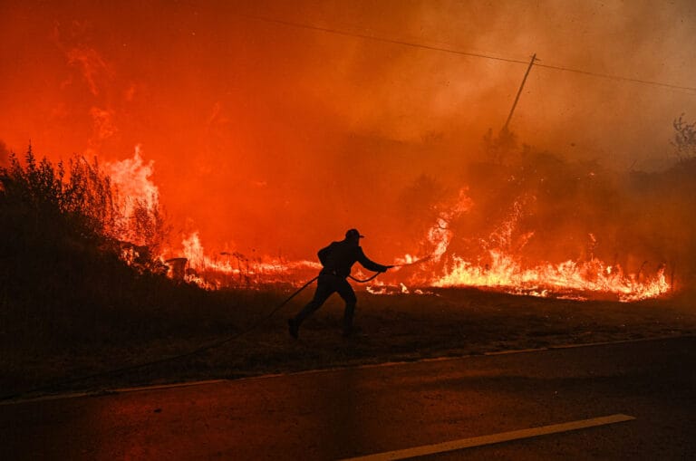 Person runs along a roadside pulling a hose toward a spreading wildfire with tall flames nearby and smoke filling the scene
