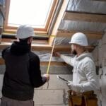 Two construction workers in hard hats measure a skylight opening in an unfinished attic using a tape measure between wooden beams