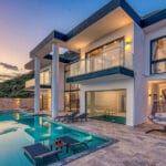 Modern villa with glass balconies overlooking a lush hillside and a sunlit infinity pool at dusk outdoor loungers scattered around