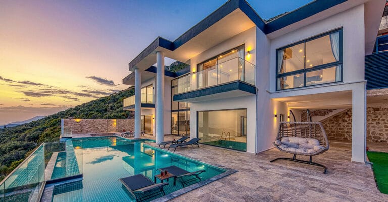 Modern villa with glass balconies overlooking a lush hillside and a sunlit infinity pool at dusk outdoor loungers scattered around