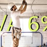 Construction worker in a white hard hat and tool belt installing wiring on an unfinished ceiling inside a renovation site