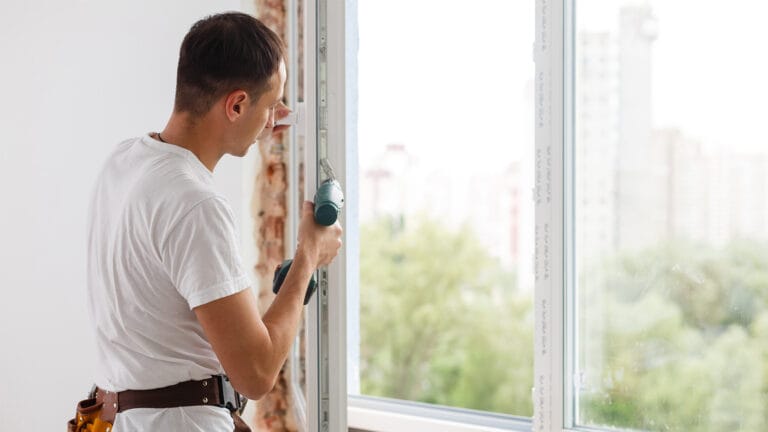 Man in a white shirt uses a cordless drill to install a window frame indoors wearing a tool belt