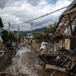 Flooded residential street with collapsed houses and scattered debris after a storm or flood event mountains in the distance and dark clouds above