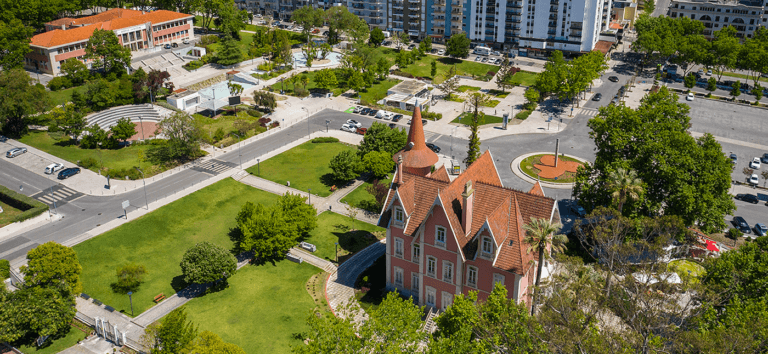Birds eye view of a pink castle like building with steep red roofs in a green city park surrounded by trees walkways and adjacent roads