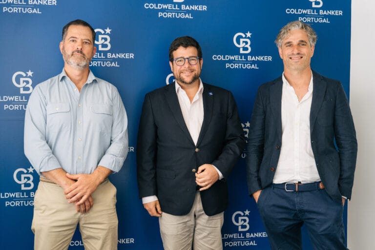 Three men in smart casualbusiness attire pose together in front of a blue backdrop branded with Coldwell Banker Portugal logos