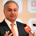 Man in a dark suit and orange tie speaks at a press conference with BPI branding in the background and microphones in front of him