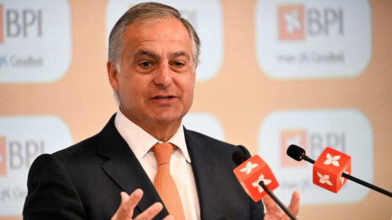Man in a dark suit and orange tie speaks at a press conference with BPI branding in the background and microphones in front of him