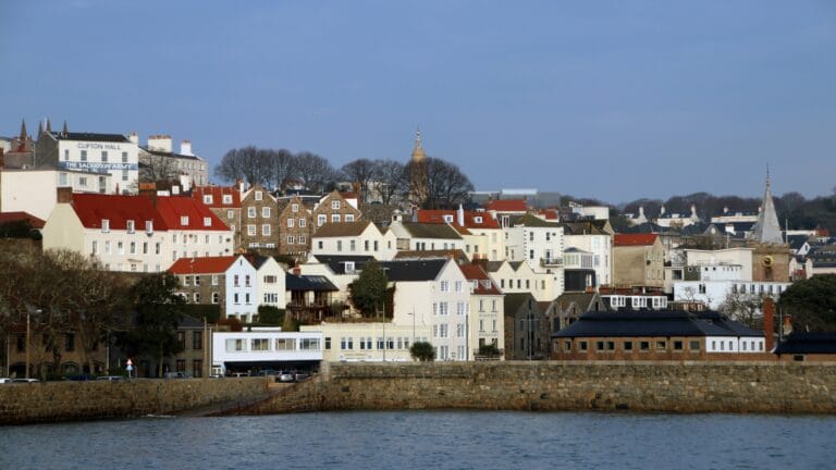 Coastal town with white and stone houses along a waterfront a stone wall and a distant church spire under a blue sky