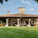 Rustic stone house with a wide covered patio and white curtains outdoor seating and a green lawn in front