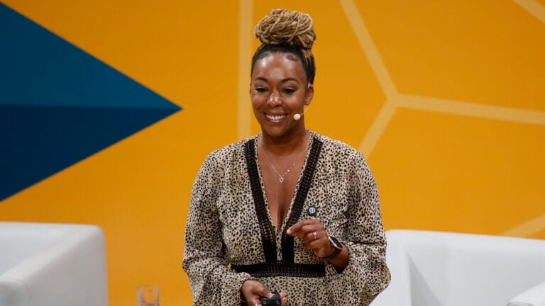 Woman presenter on stage wearing a leopard print dress with a headset microphone smiling against an orange geometric backdrop