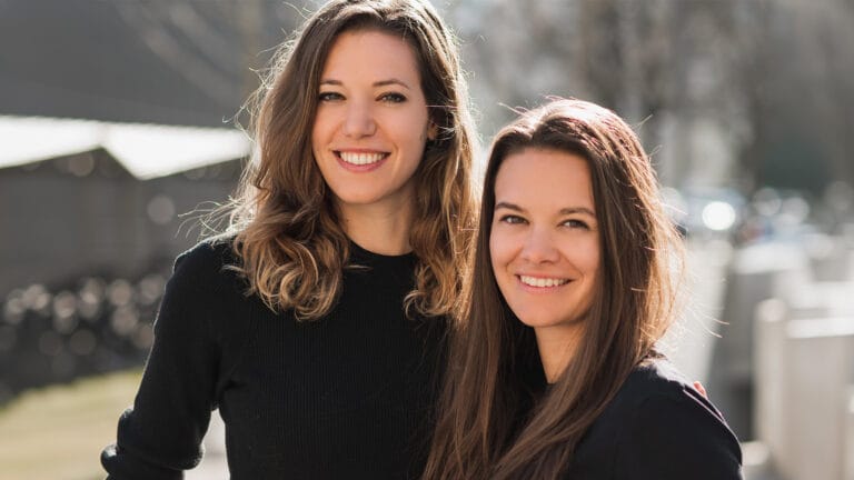 Two women smiling and posing outdoors on a sunny day standing close together in black tops
