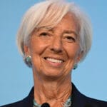 Smiling older woman with short white hair turquoise earrings and necklace wearing a dark blazer against a blue background