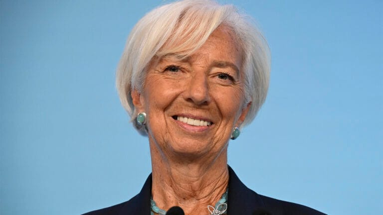 Smiling older woman with short white hair turquoise earrings and necklace wearing a dark blazer against a blue background
