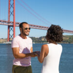 Man wearing a pink t shirt and sunglasses smiles at a woman in a white dress by the water with a large red suspension bridge in the background