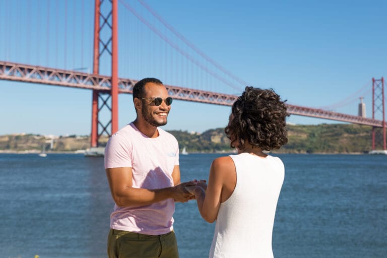Man wearing a pink t shirt and sunglasses smiles at a woman in a white dress by the water with a large red suspension bridge in the background