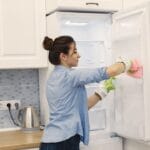 Woman wearing gloves cleans the inside of an open refrigerator with colorful cloths in a bright kitchen