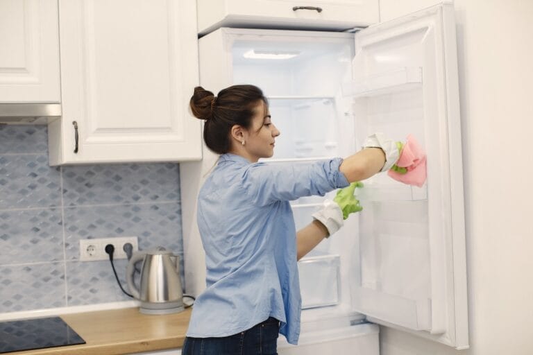 Woman wearing gloves cleans the inside of an open refrigerator with colorful cloths in a bright kitchen