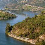 A train winds along a curved track built into a green hillside beside a calm blue lake with villages on the far shore