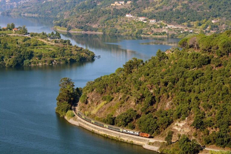 A train winds along a curved track built into a green hillside beside a calm blue lake with villages on the far shore