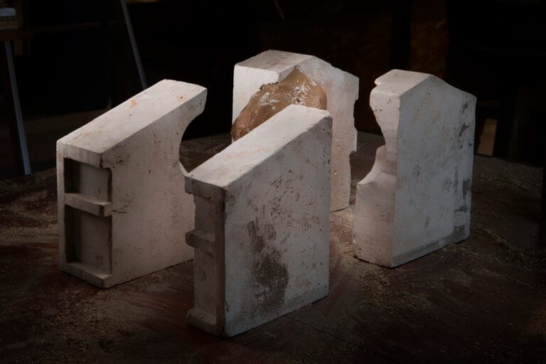 Four pale concrete blocks arranged in a rough circle on a dusty workshop floor with a brown clay like lump in the center