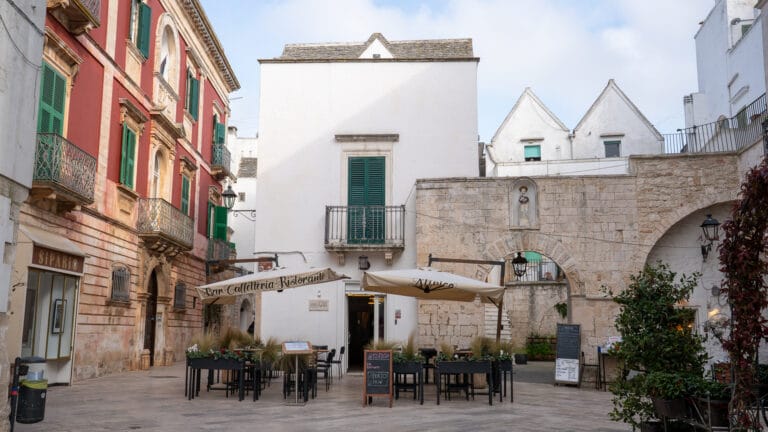 Stone paved town square with outdoor cafe seating under beige canopies flanked by a white building in the center and a red brick building with green shutters on the left plus a stone arch on the right