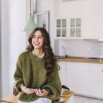 Smiling woman in a green knit sweater sits at a kitchen island holding a smartphone