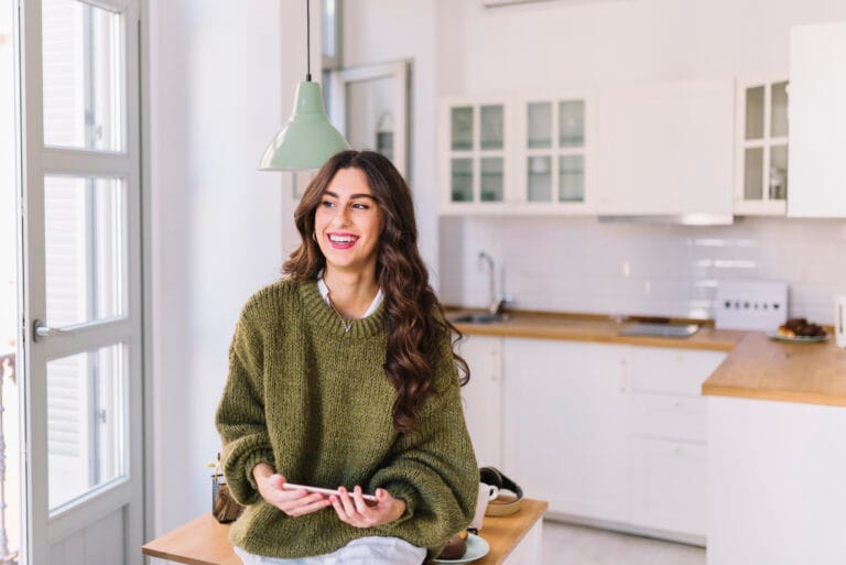 Smiling woman in a green knit sweater sits at a kitchen island holding a smartphone