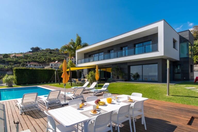 Modern two story white and gray villa with glass balcony overlooking a sunny poolside dining area and yellow umbrellas on a wooden deck