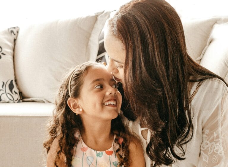 Mother with long dark hair leans in to kiss a smiling young girl on the nose They sit close on a light couch sharing a loving moment
