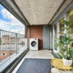 A glass walled balcony with a washing machine against a brick wall overlooking a cityscape of red brick buildings and blue sky