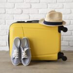 Yellow hard shell suitcase with a straw hat on top and gray sneakers resting beside it on a wooden floor against a white brick wall